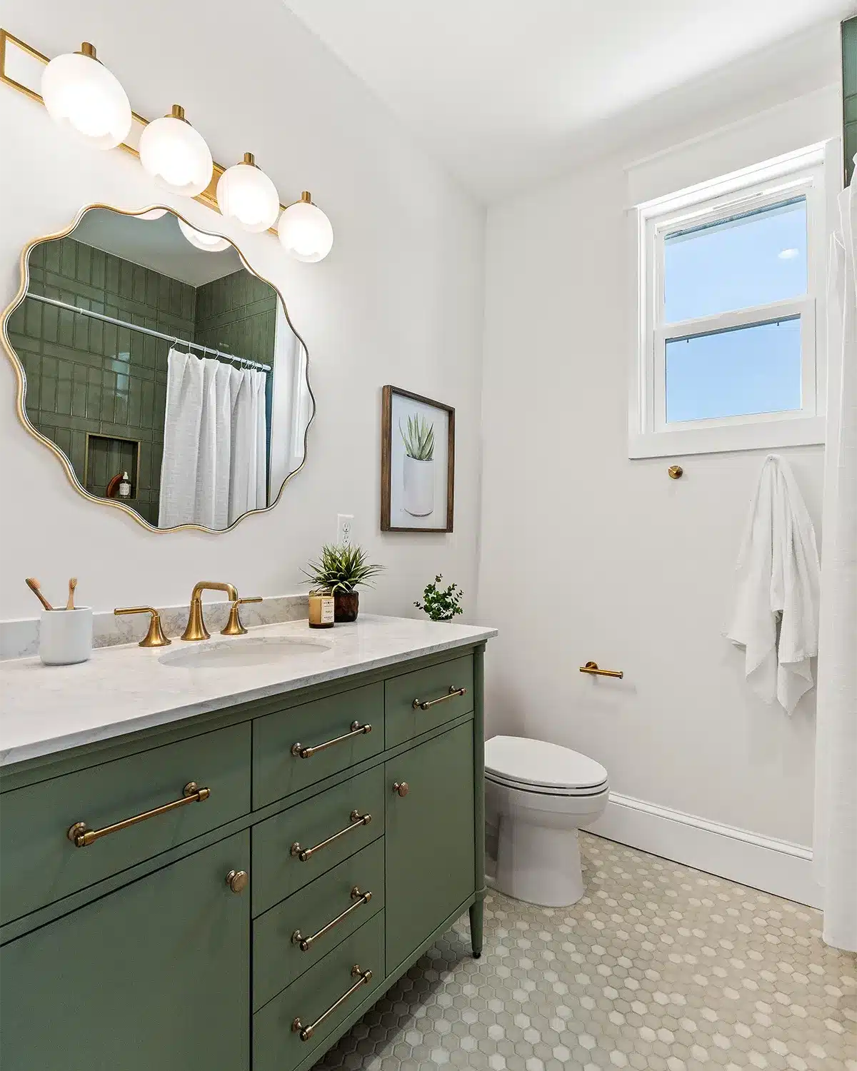 A small bathroom features a large round mirror, light colors, sage vanity, and hexagonal floor tile.