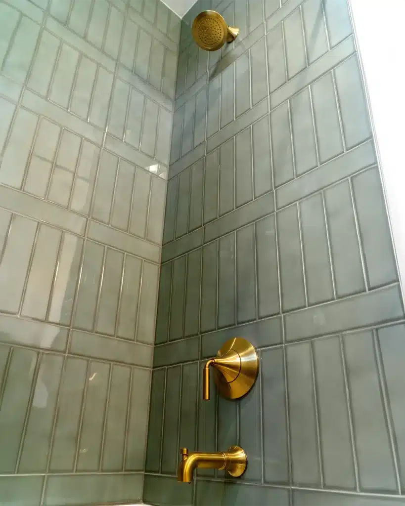 Long vertical, polished, sage green tiles extend up the walls of a shower featuring a gold shower head and faucet.
